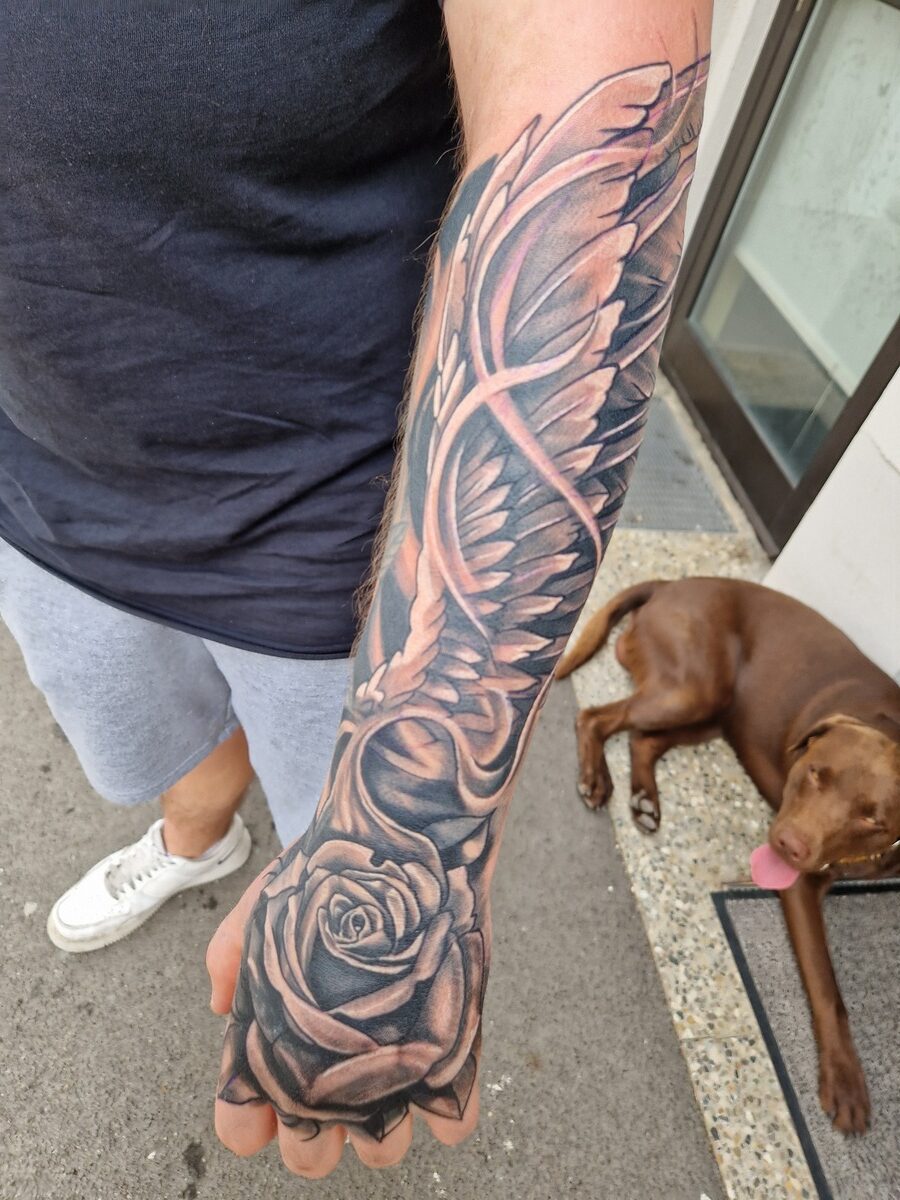 Mann mit schwarzem Oberteil, tätowierter Arm mit Rosen und Flügeln, Hund liegt im Hintergrund.