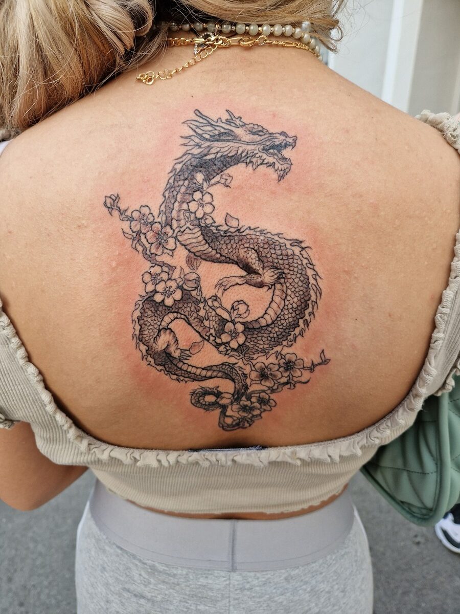 Frau mit blonden Haaren, trägt ein Top; Drache-Tattoo auf Rücken, mit Blumen, schwarzer Tinte.