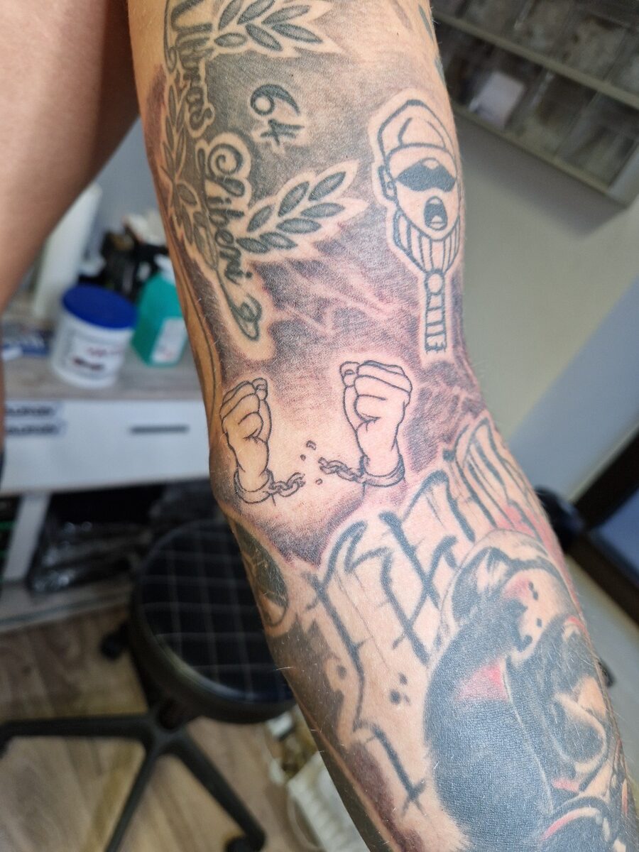 Unterarm-Tattoo mit Händen in Fesseln, Schriftzug und Karikaturfigur, bunte Details.