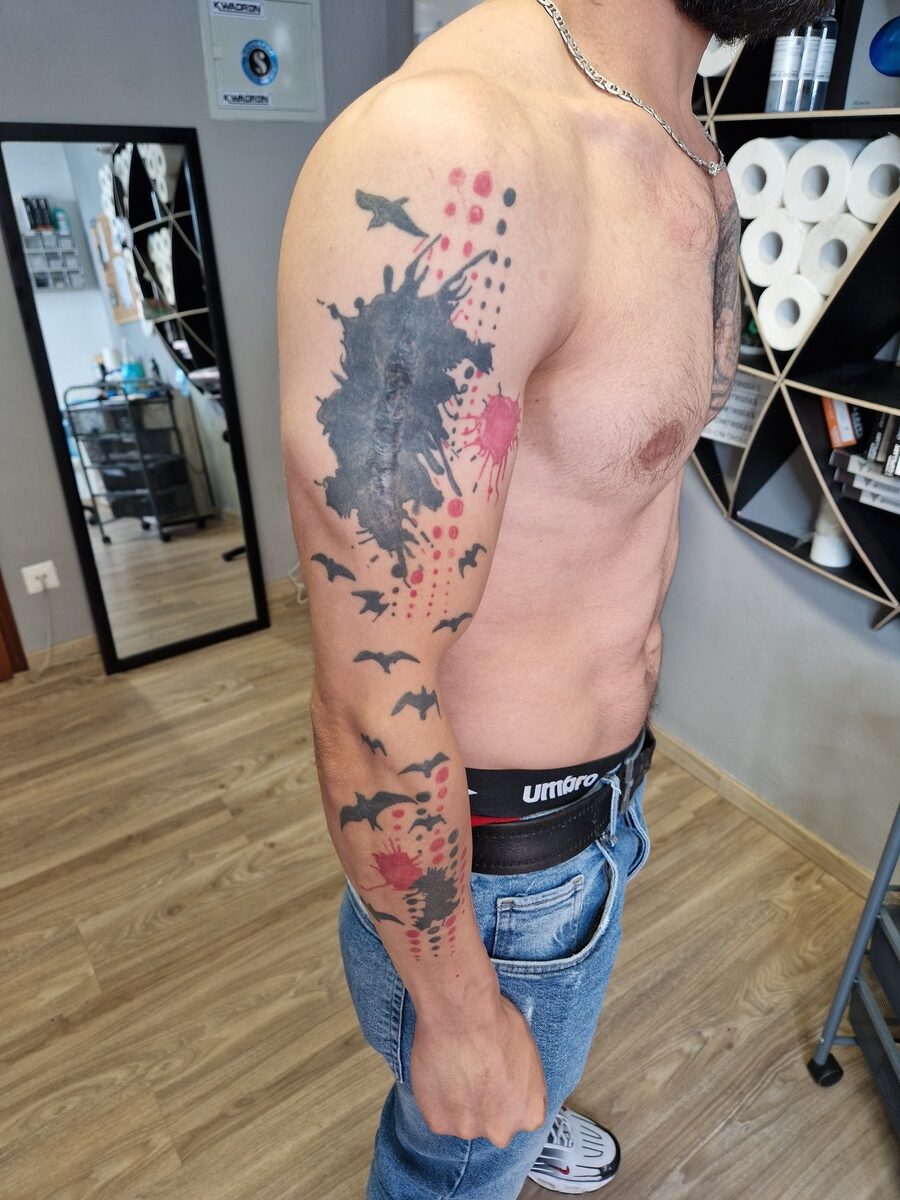 Ein Mann mit Bart zeigt farbige Tattoos auf dem Arm; er trägt Jeans, steht neben Regalen.