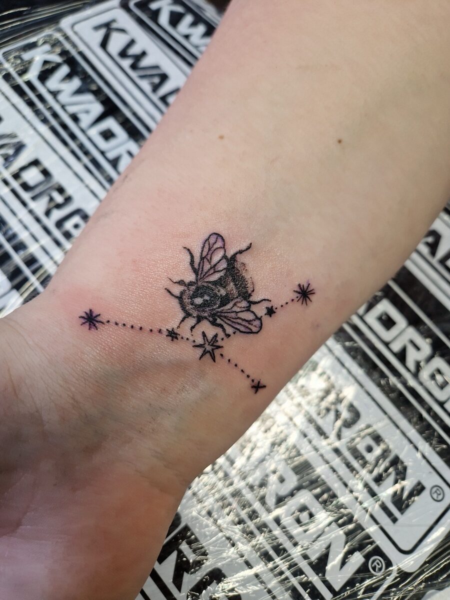 Tätowiertes Handgelenk mit Bienenmotiv und Sternen, schwarzer Tinte, zarte Linien.