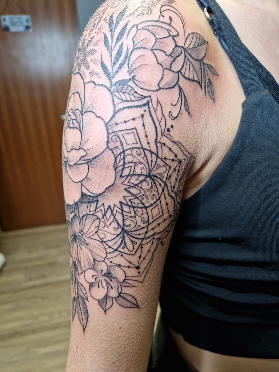 Frau mit schwarzem Oberteil zeigt großes florales Tattoo auf dem Oberarm, detailliert und komplex.