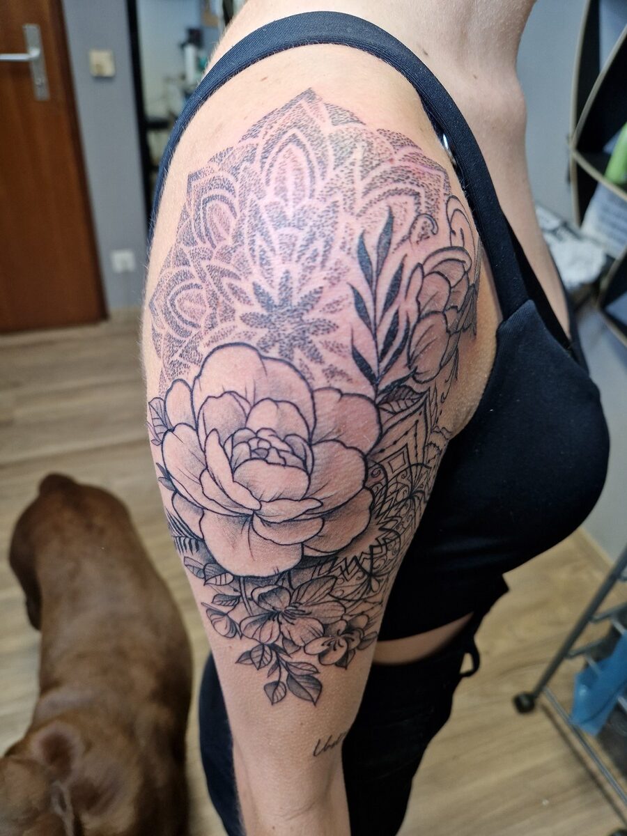 Person mit floralen Tattoos auf dem Oberarm, trägt schwarzes Top, Hund im Hintergrund.