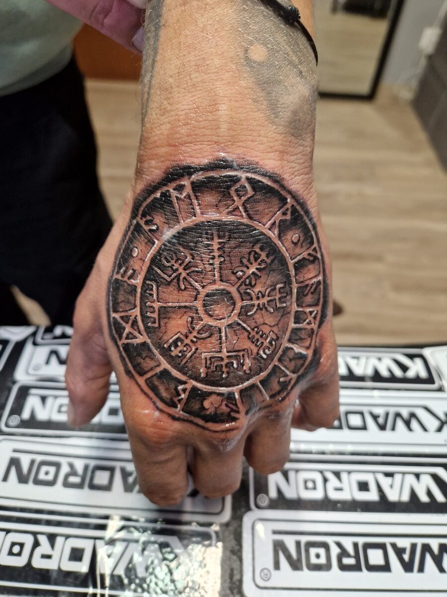 Tätowierte Hand mit aztekischem Design, dunkelgrau-schwarz, auf Holztisch.