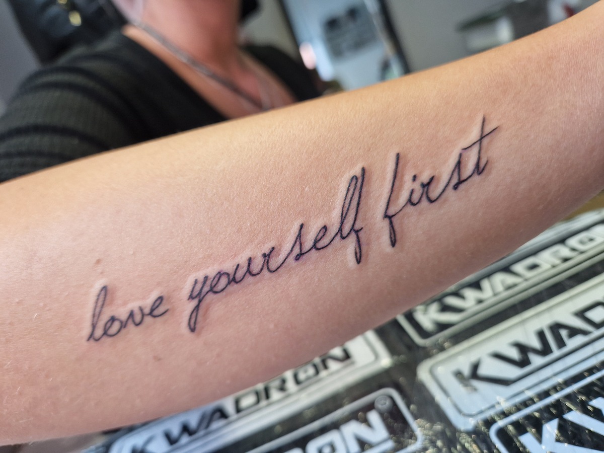 Nahaufnahme eines Arms mit schwarzem Schriftzug-Tattoo "love yourself first".