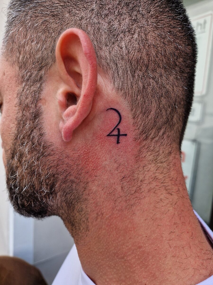 Nahaufnahme eines Mannes mit kurzem Haar und einem Tattoo hinter dem Ohr in Form von "24".