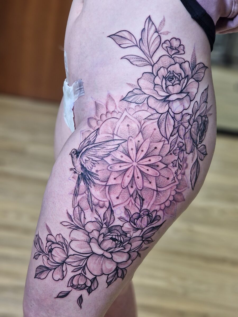 Tattoo am Bein mit Blumen, Vogel und Mandala; schwarz-grau, komplexe Details, Pflaster sichtbar.
