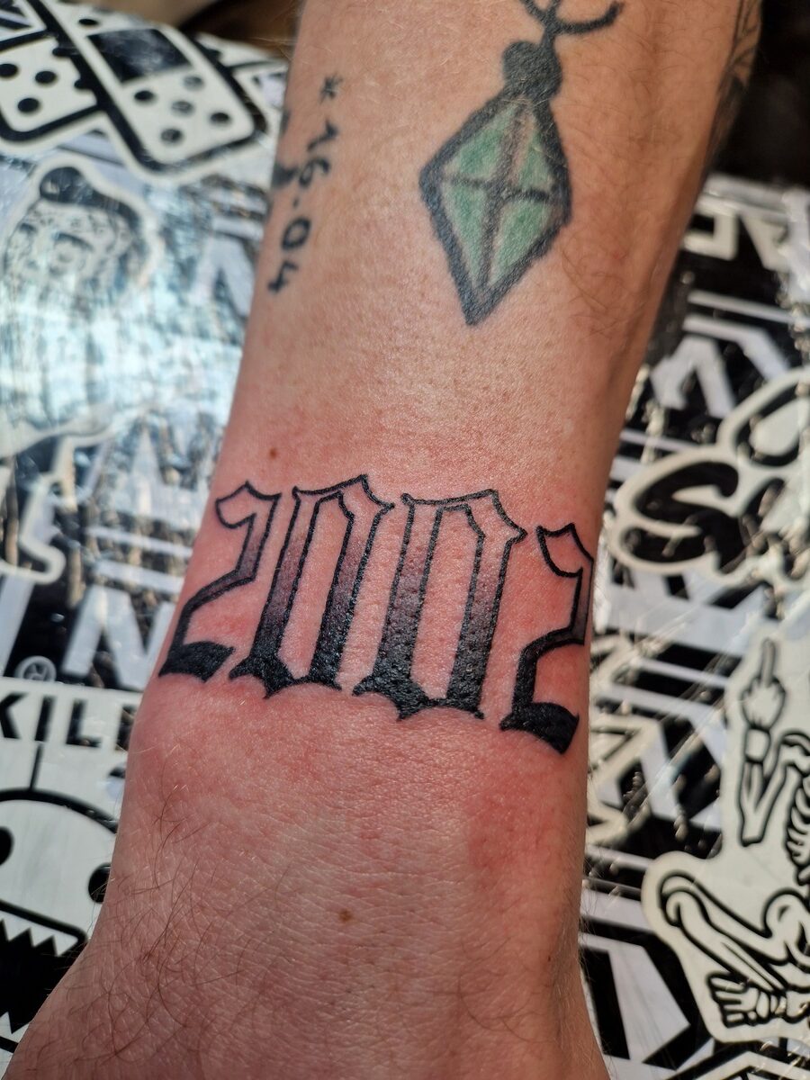 Arm mit "2002"-Tattoo und grüner Diamant-Tätowierung auf bunter Unterlage.