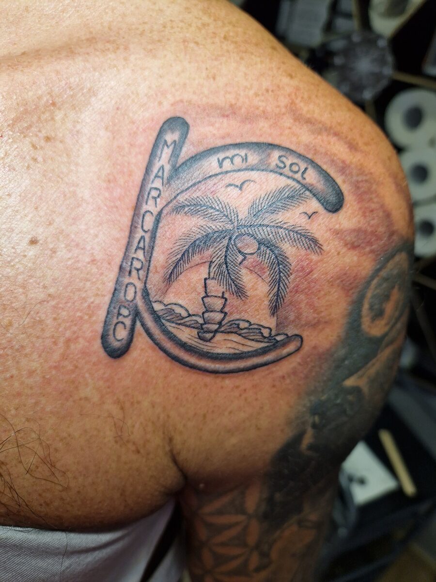 Tattoo auf einer Schulter: Palmen, Sonnenuntergang, Schrift "MARCAR OPC", "Sol".