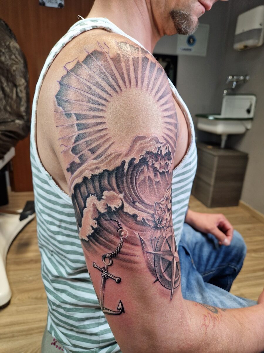 Mann mit gestreiftem Hemd zeigt Arm-Tattoo: Sonne, Wellen, Anker, Windrose; sitzend, entspannt.