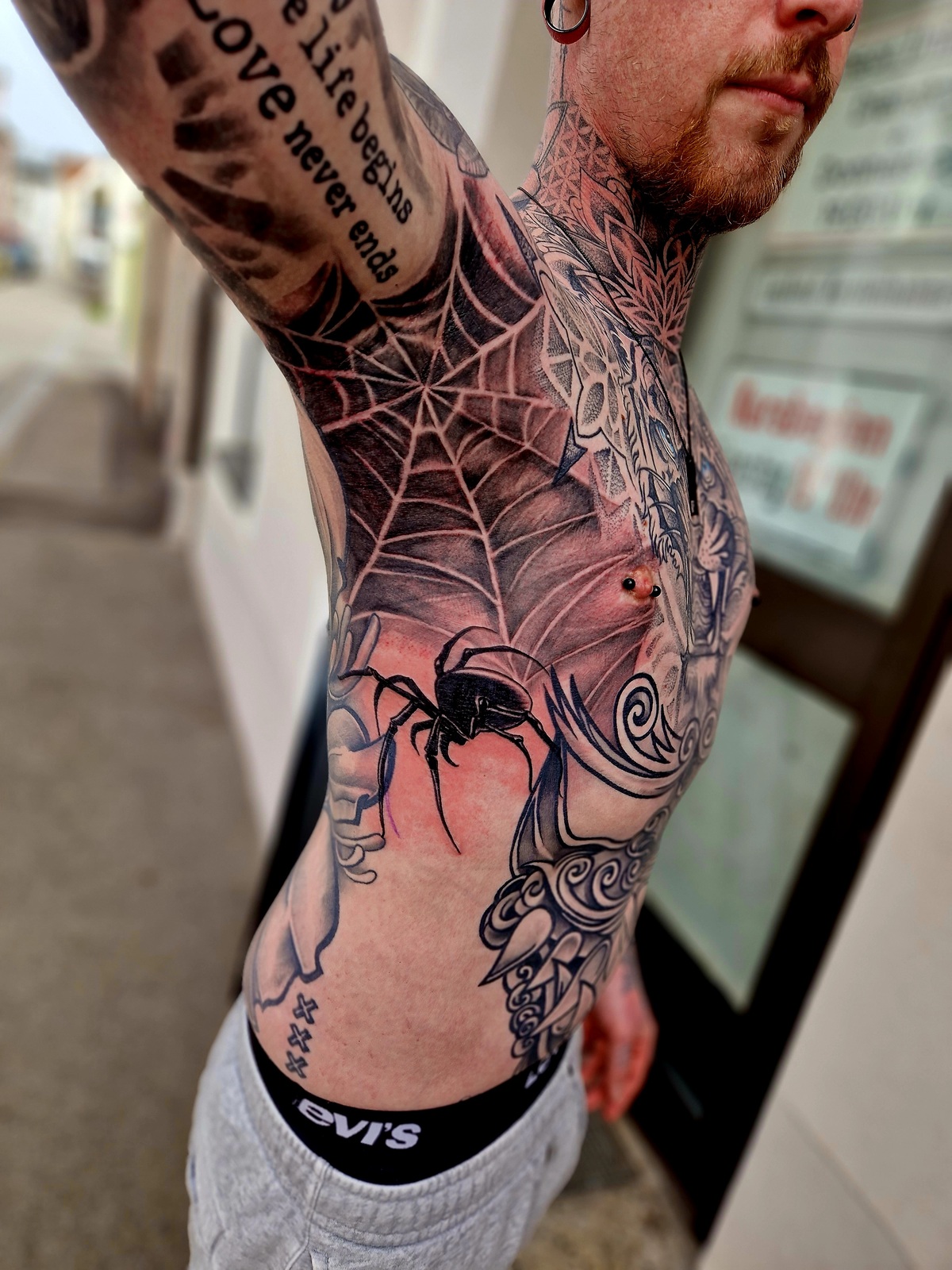 Mann mit Tattoos am Oberkörper, Spinnennetz-Design, stechender Blick, schwarz-weiße Muster.