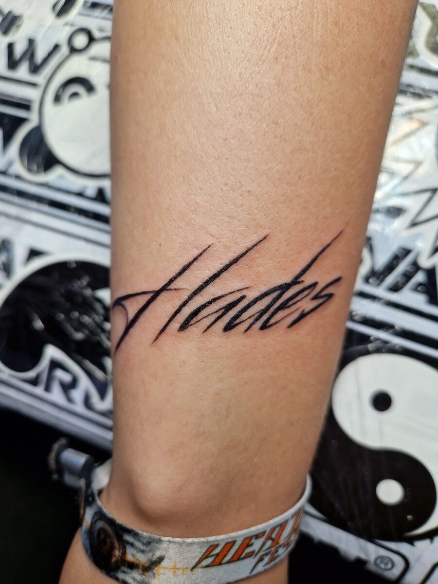 Ein Arm mit einem Tattoo des Wortes "Hades" in schwarzer Tinte, Yin-Yang-Hintergrund.