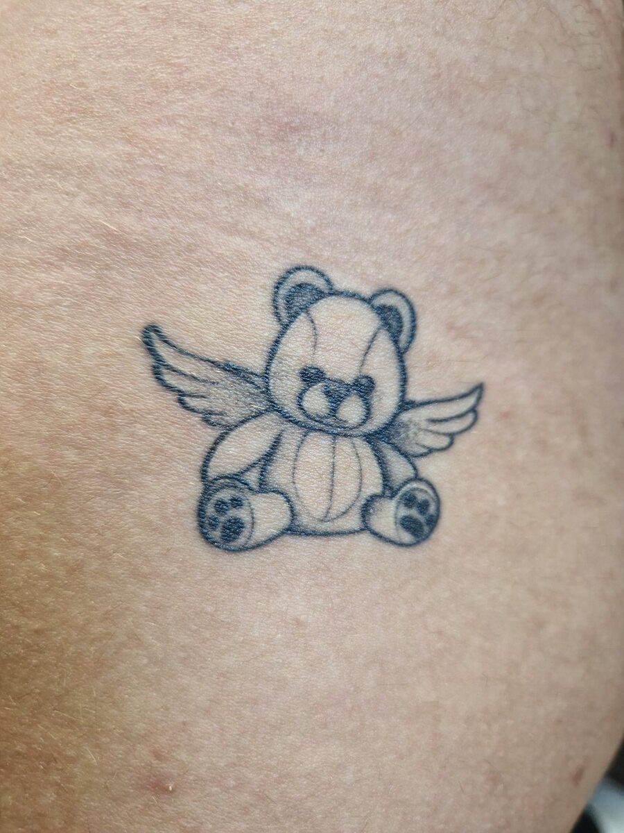 Tattoo eines Teddybären mit Flügeln auf heller Haut; blauer Umriss; niedliches Design.